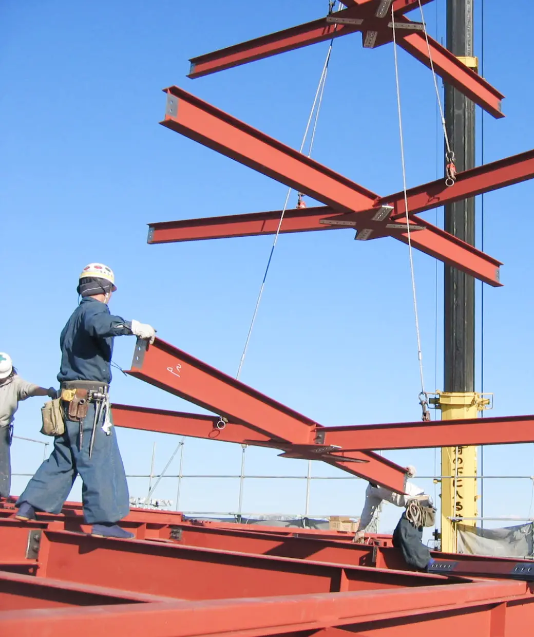 建設現場で、赤い鉄骨を組み立てる作業員がクレーンを使用して作業している様子