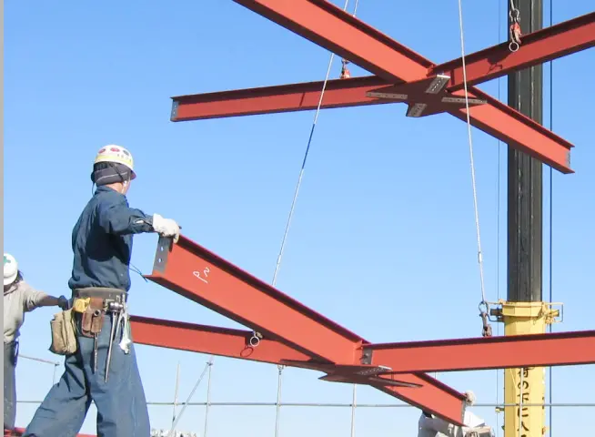 建設現場で、赤い鉄骨を組み立てる作業員がクレーンを使用して作業している様子